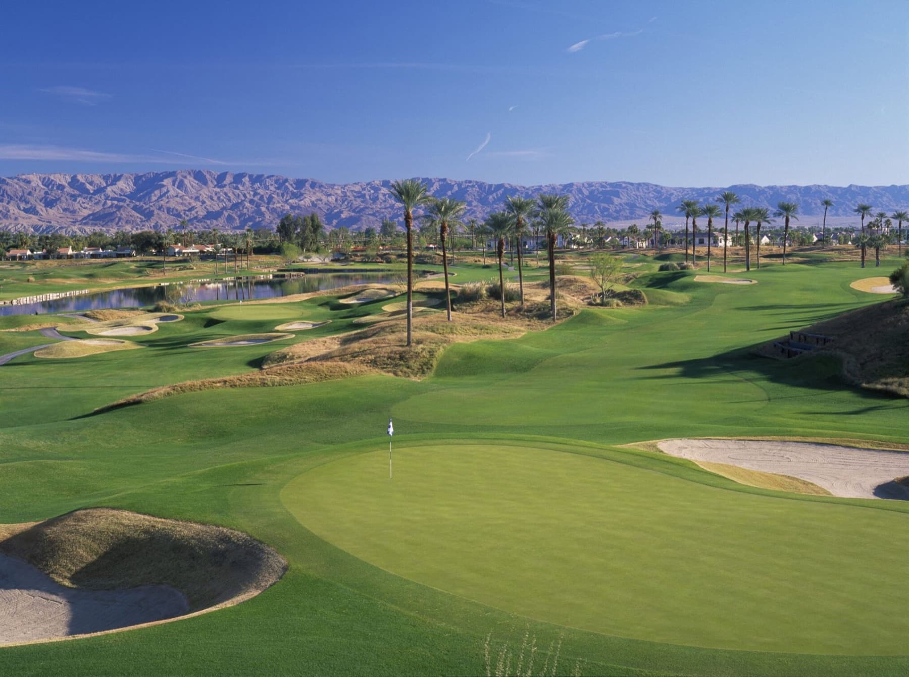 A Dunes Course green ringed by bunkers, water and palms below the desert mountains