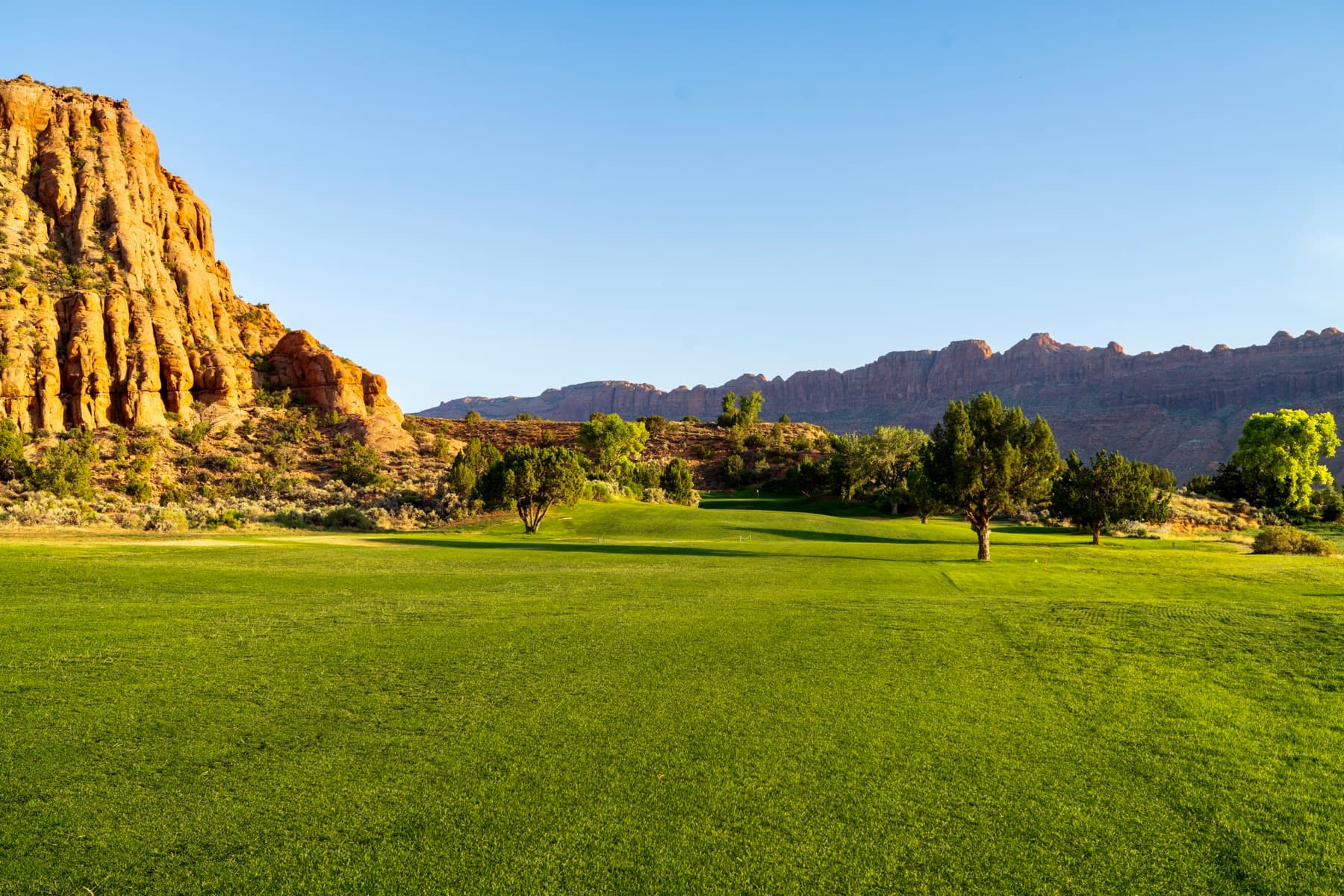 A fairway running up toward the green beneath a towering sandstone cliff, red-rock ridgelines beyond